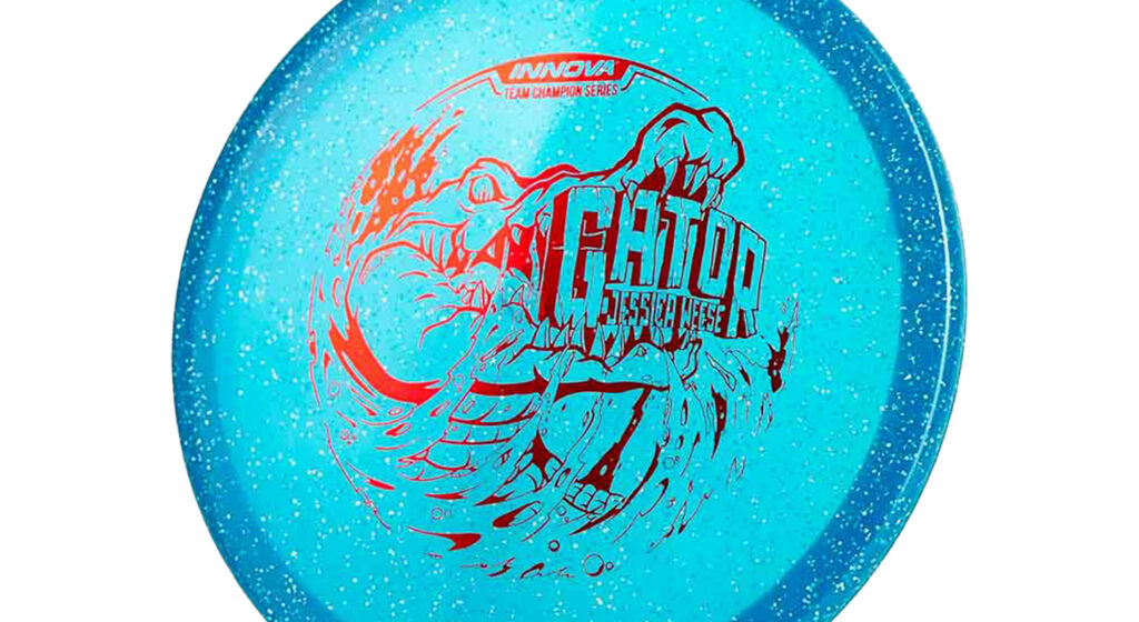 An Innova Metal Flake Gator (Jessica Weese 2022 Tour Series) with Blue metallic and glittery color and red stamp. The image depicted on the disc features an alligator poised to bite into the text, with a dynamic water splash surrounding it.